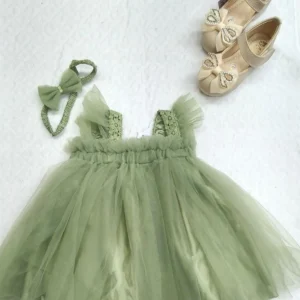 A sage green baby girl tulle dress with lace straps, matching headband, and beige shoes with pearl bows arranged on a white background.